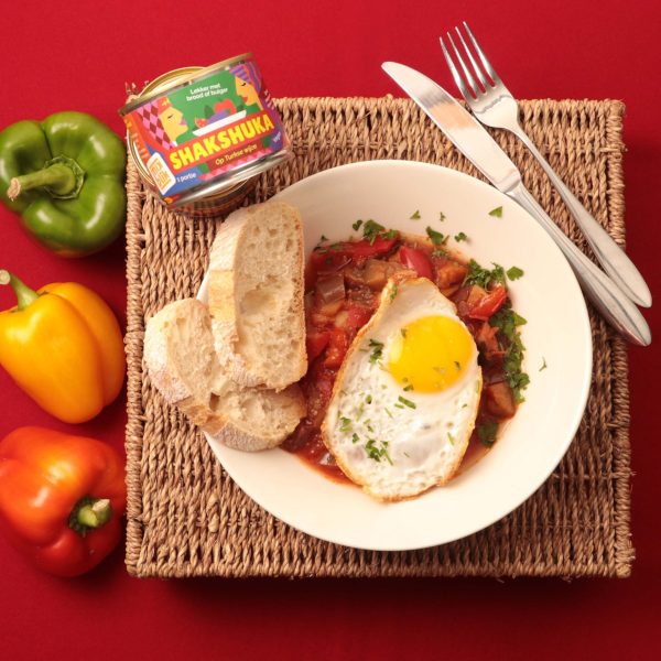 Mixblik klein shakshuka blik naast een shakshuka gerecht met ei en brood op tafel. Groene, gele en rode peper op tafel.